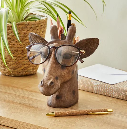 Photo of a small wooden head created in the shape and features of a giraffe. The giraffe has a slot in the top of it's head where a pair of glasses can be rested to hold them. 